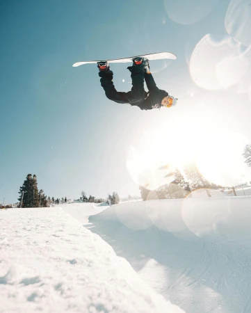 A snowboarder performs an inverted aerial trick high above a snowy slope on a bright, sunny day in an outdoor winter setting.