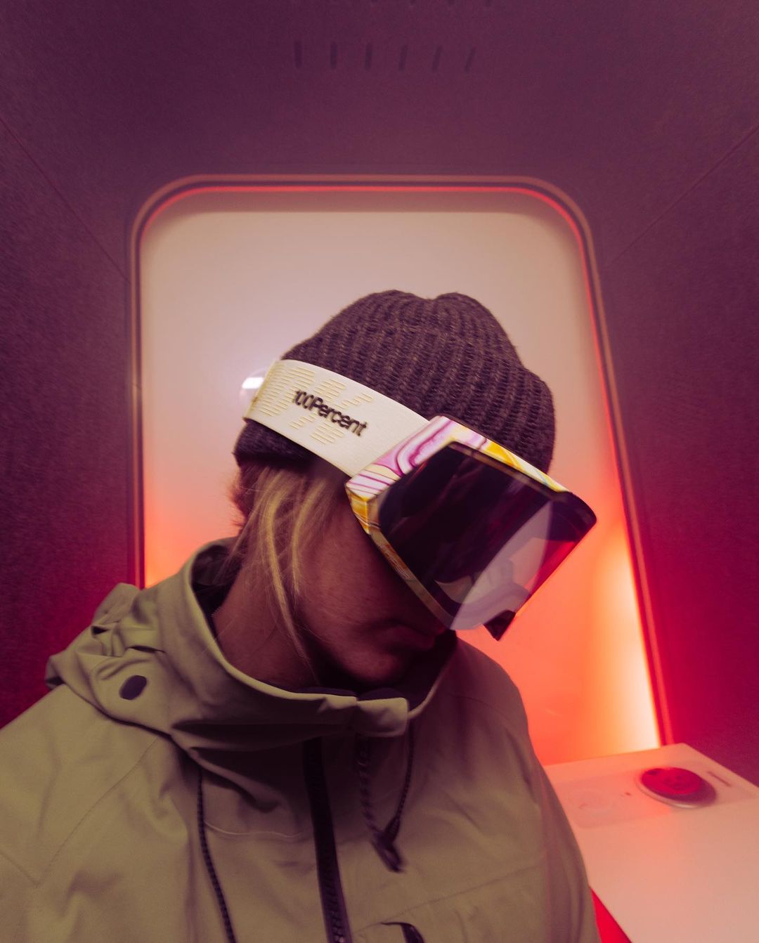 Person wearing beanie and ski goggles indoors with white wall and orange accent lighting, close-up shot showing head and shoulders.