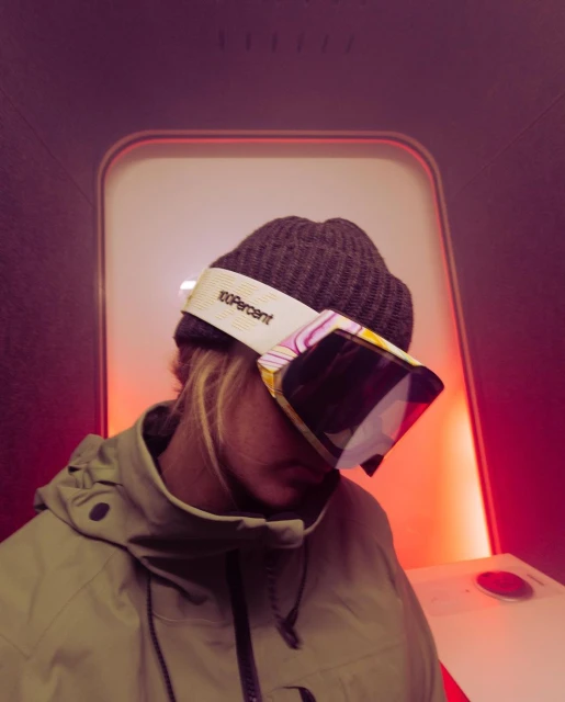 Person wearing beanie and ski goggles indoors with white wall and orange accent lighting, close-up shot showing head and shoulders.