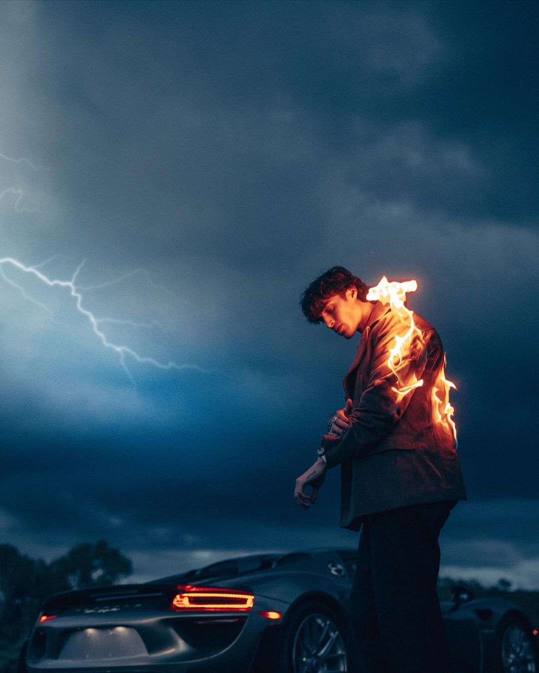 Artistic portrait of a male model in jacket with fire effect, standing in front of a silver sports car and stormy sky at night.