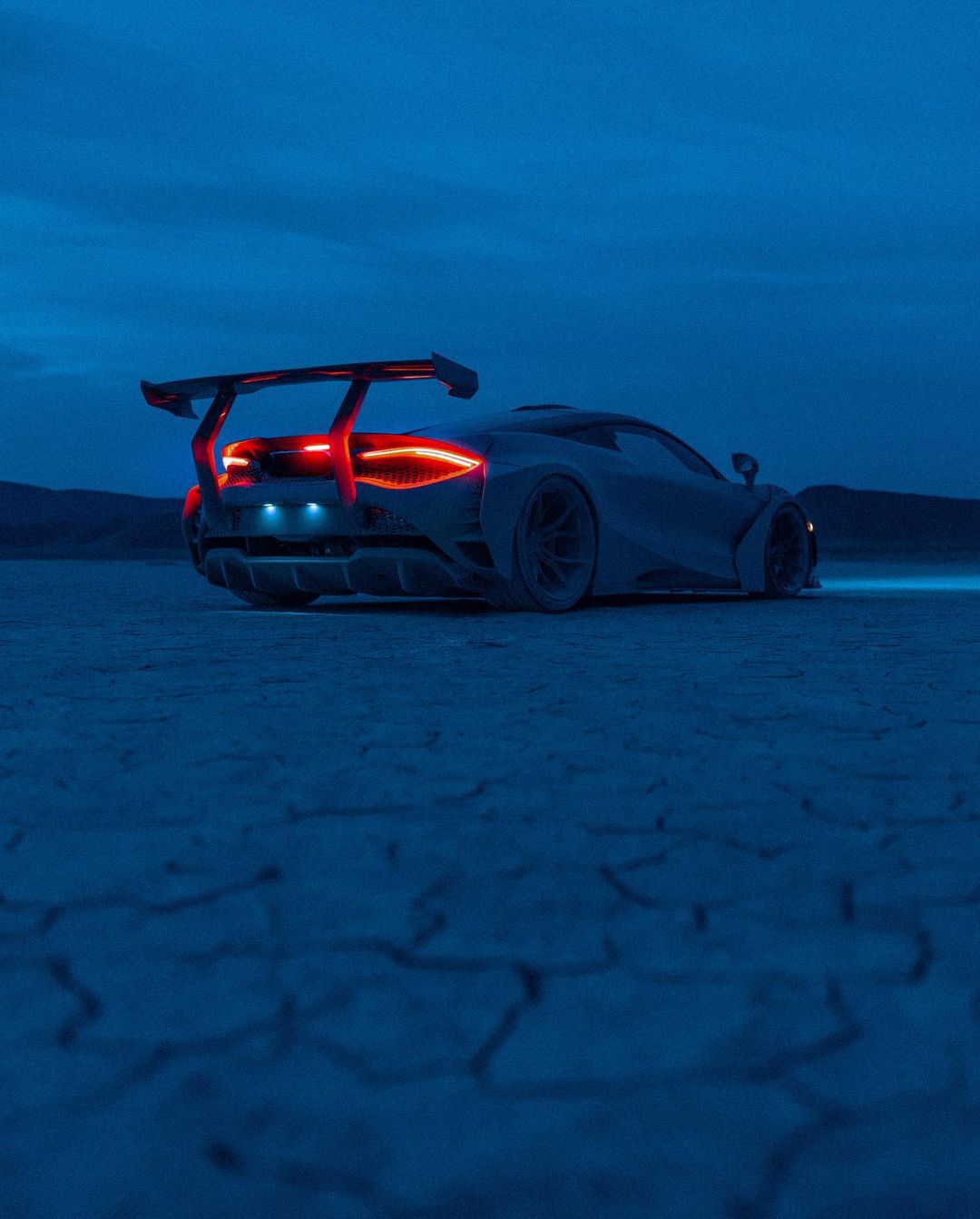 Sleek silver sportscar with a rear spoiler parked on a cracked dry lakebed under a dusk sky, taillights glowing red, artistic render.