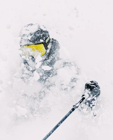 A person in ski gear, wearing a helmet and goggles, is seen from a low angle while skiing in deep, fresh powder snow.