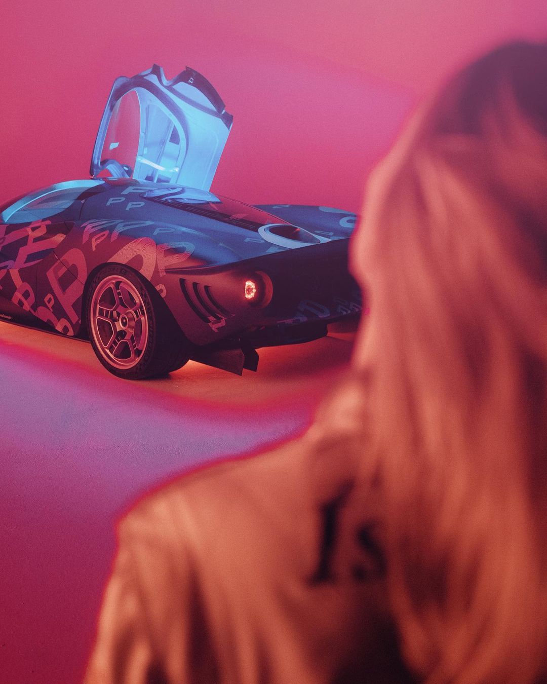 A stylized sports car with its doors open is parked against a pink background, with a person standing in the foreground.