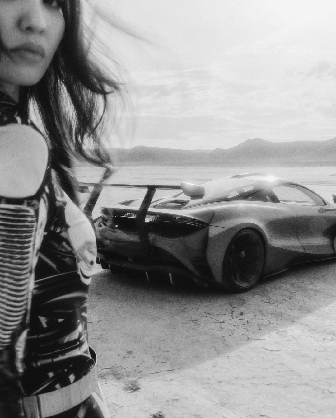 Monochrome shot of an Asian woman in a high-fashion latex suit with a sportscar on a cracked-earth landscape, high-contrast and dramatic.