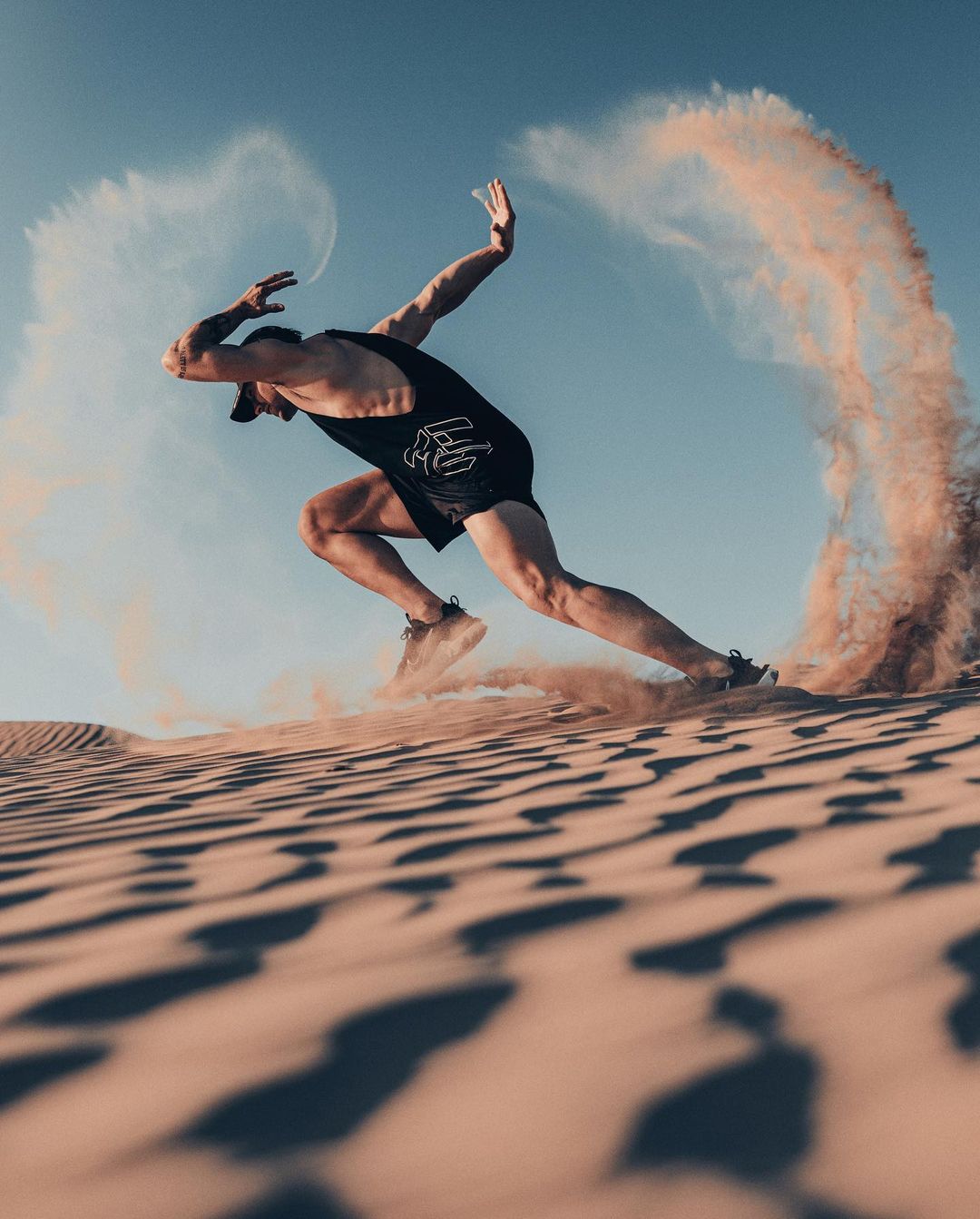 A man sprints on a sand dune with sand billowing, wearing a black athletic outfit, capturing a moment of intense desert fitness.