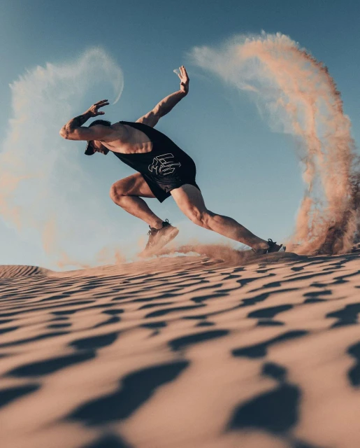 A man sprints on a sand dune with sand billowing, wearing a black athletic outfit, capturing a moment of intense desert fitness.
