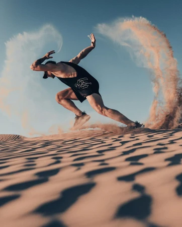A man sprints on a sand dune with sand billowing, wearing a black athletic outfit, capturing a moment of intense desert fitness.
