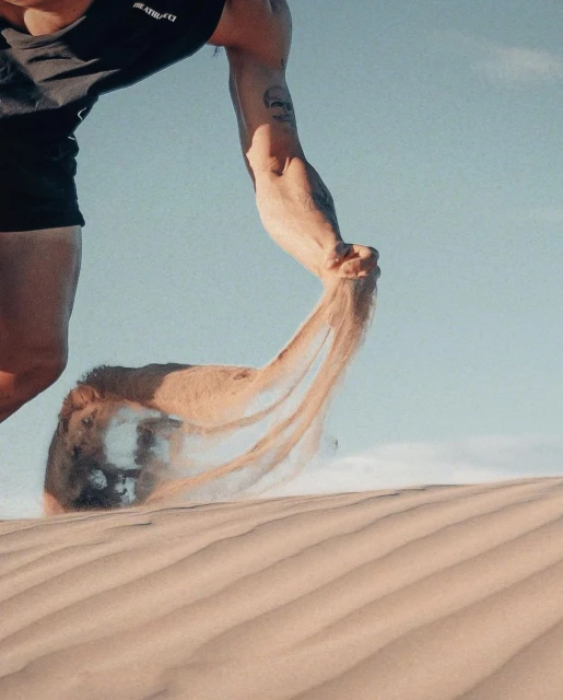 Adult male pours sand through his fingers on a bright, sunny day at the beach with blue skies, wearing a black athletic top.