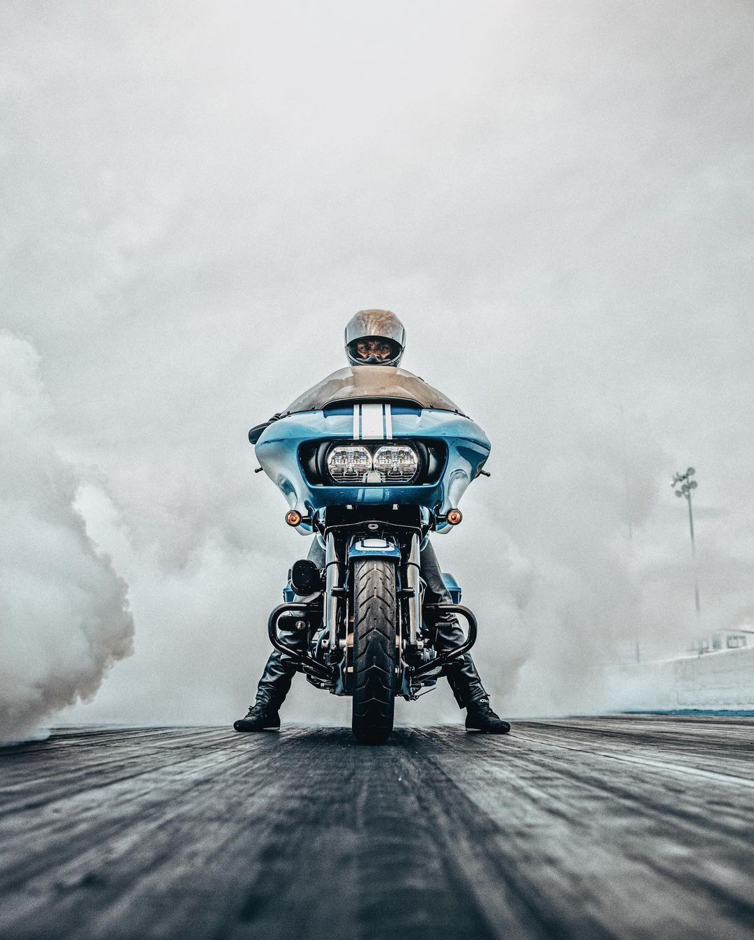 Motorcyclist on a blue motorcycle performs a burnout on a track surrounded by smoke with overcast skies above and a low camera angle.