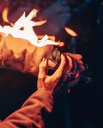 Close-up of hands wearing rings and a luxury wristwatch as bright flames flicker nearby in warm ambient light.