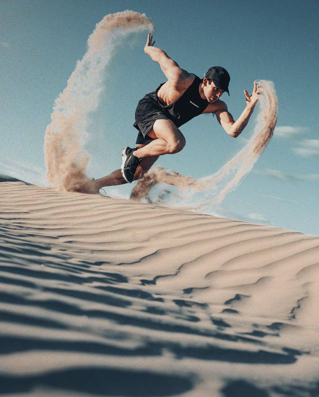Athletic man in activewear powerfully leaps across a sand dune, kicking up a swirl of sand on a bright day with a clear blue sky.