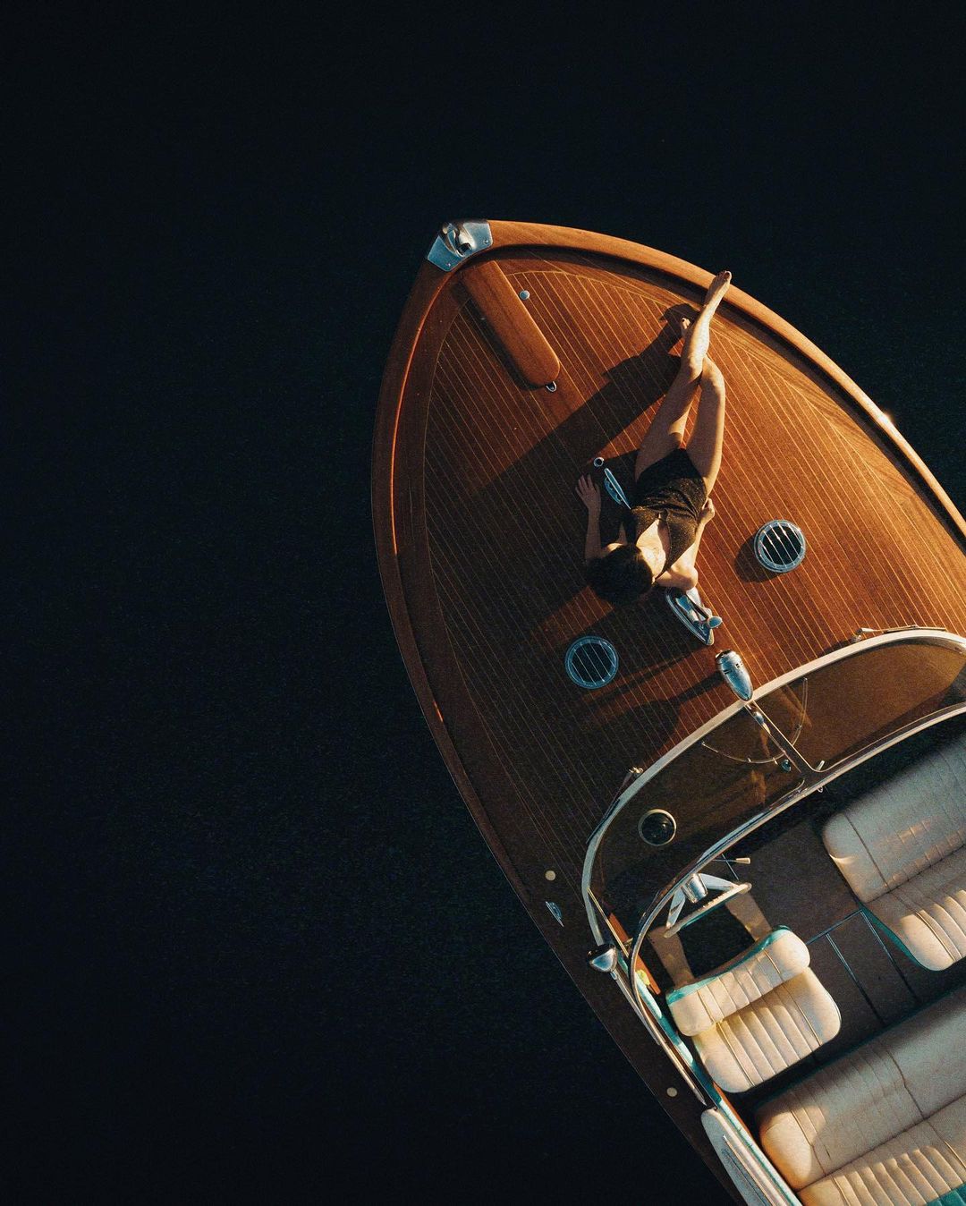 Aerial view of a person lying on the wooden deck of a motorboat floating on dark water, showcasing luxury and leisure.