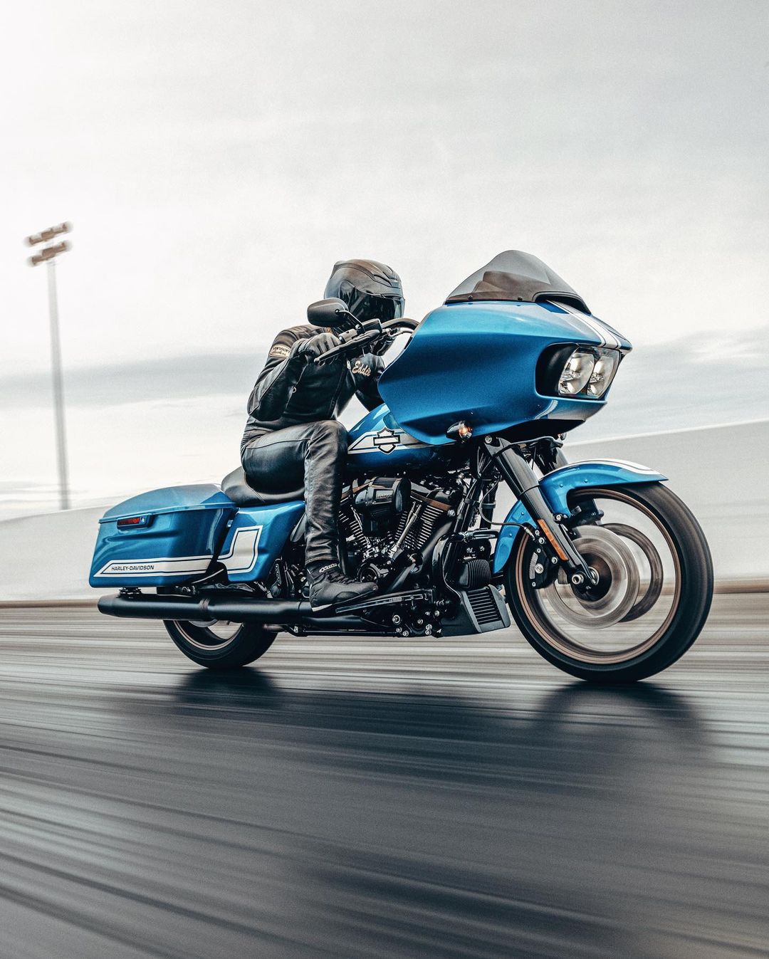 A person wearing a black helmet and leather jacket rides a blue Harley Davidson motorcycle on an asphalt track under an overcast sky.
