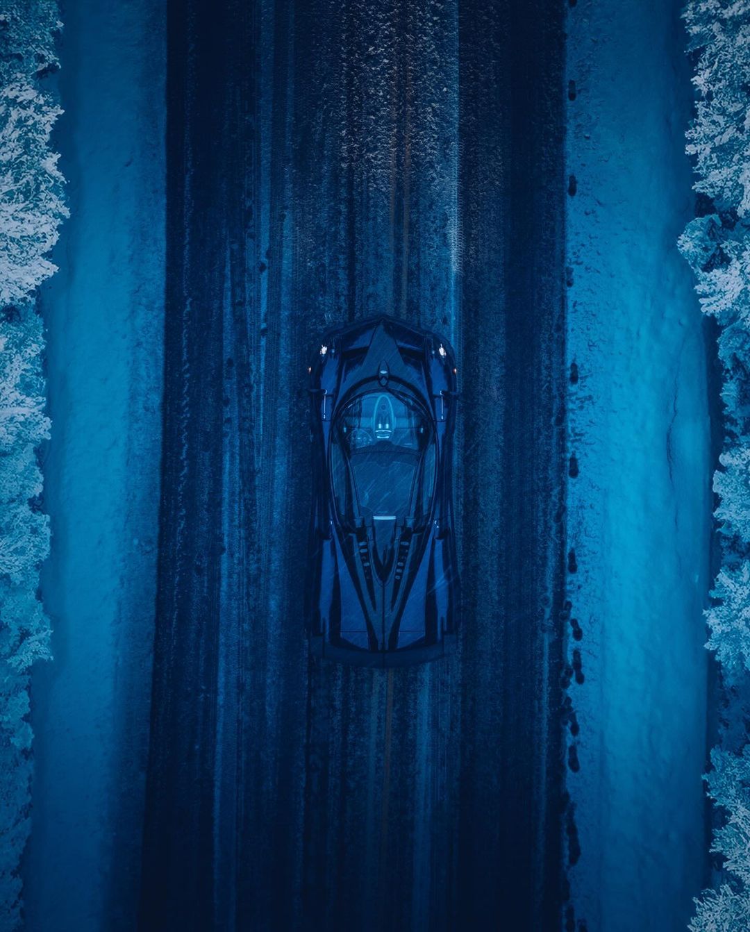Overhead shot of a dark blue sports car driving on a snow-covered road through a forest in winter, creating a moody automotive scene.