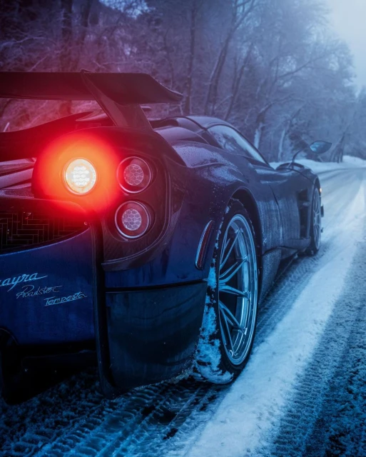 Rear view of a blue Pagani sports car on a snowy road with illuminated taillights and surrounding snow-covered trees on a cold day.
