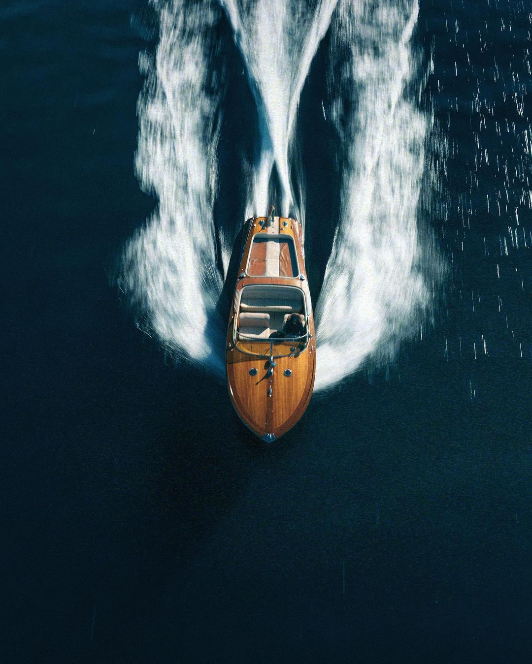 Aerial view of a classic wooden speedboat gliding on the water, leaving a trail of white water in its wake, sunny day.