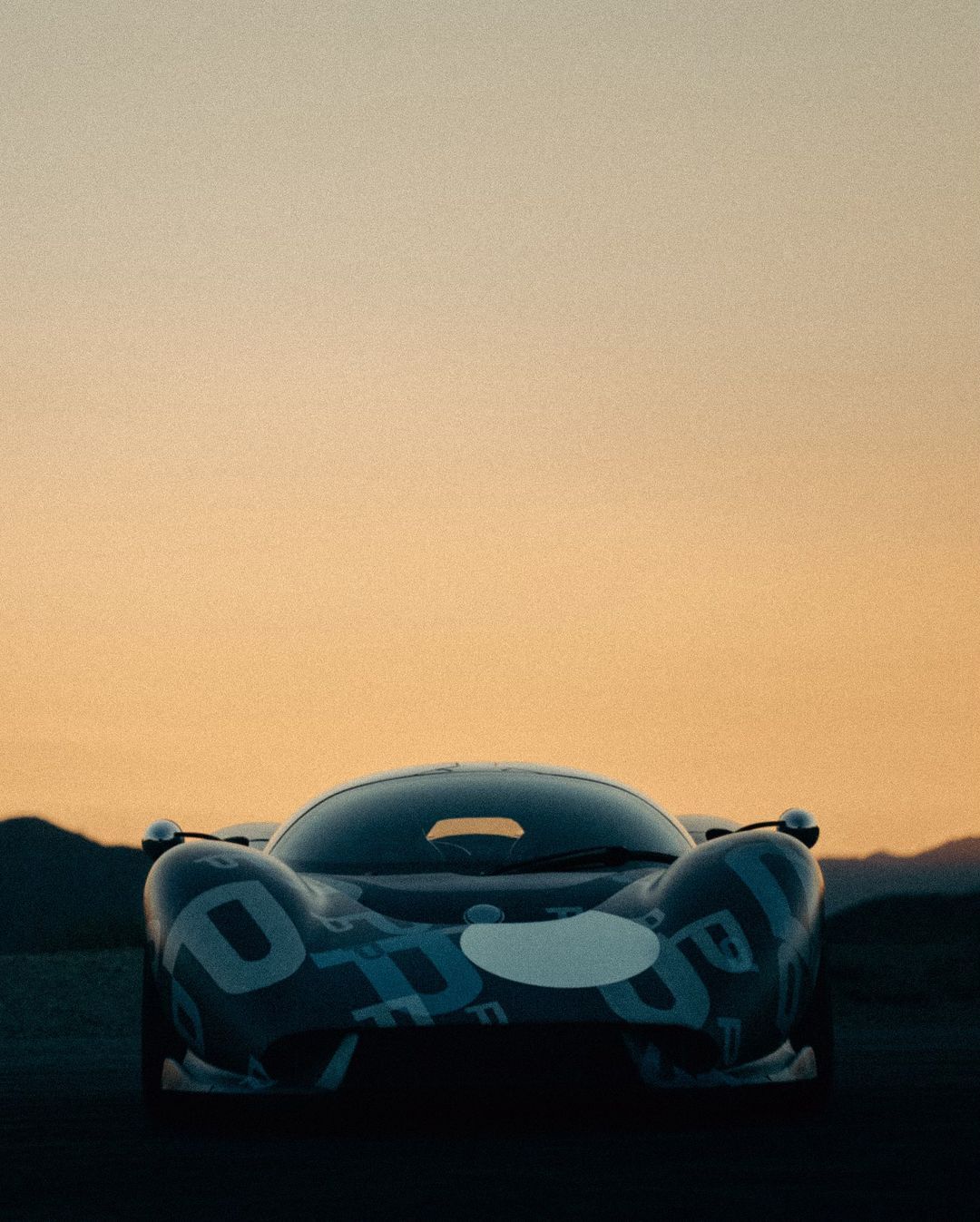 Front view of a blue Porsche race car with white decals, set against a background of mountains under an orange sky at dusk.