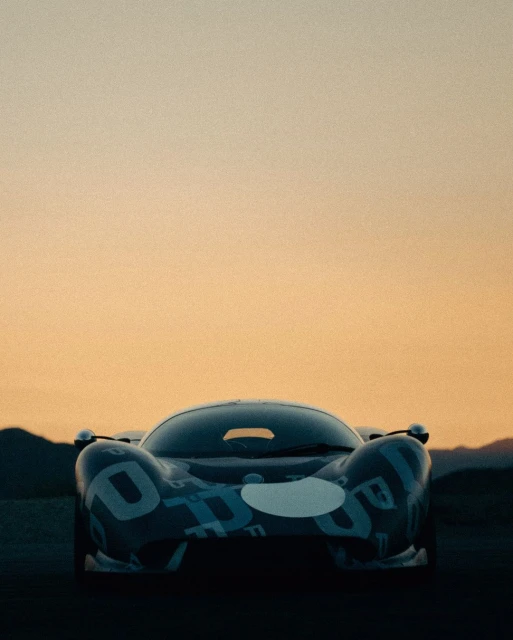 Front view of a blue Porsche race car with white decals, set against a background of mountains under an orange sky at dusk.