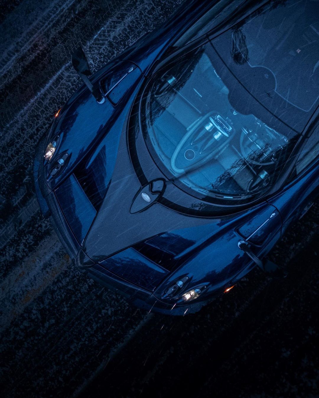 Overhead shot of a sleek, dark blue Pagani Zonda hypercar with its headlights on, sitting on dark pavement. High-performance automotive design.