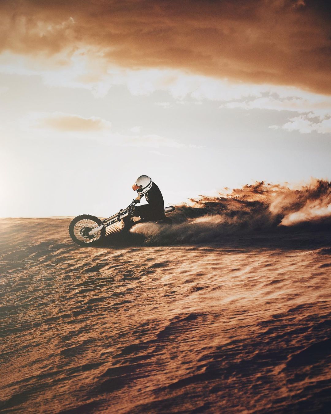 A person in a helmet rides a motocross bike across a brown, sandy landscape under a cloudy sky, kicking up sand behind them.