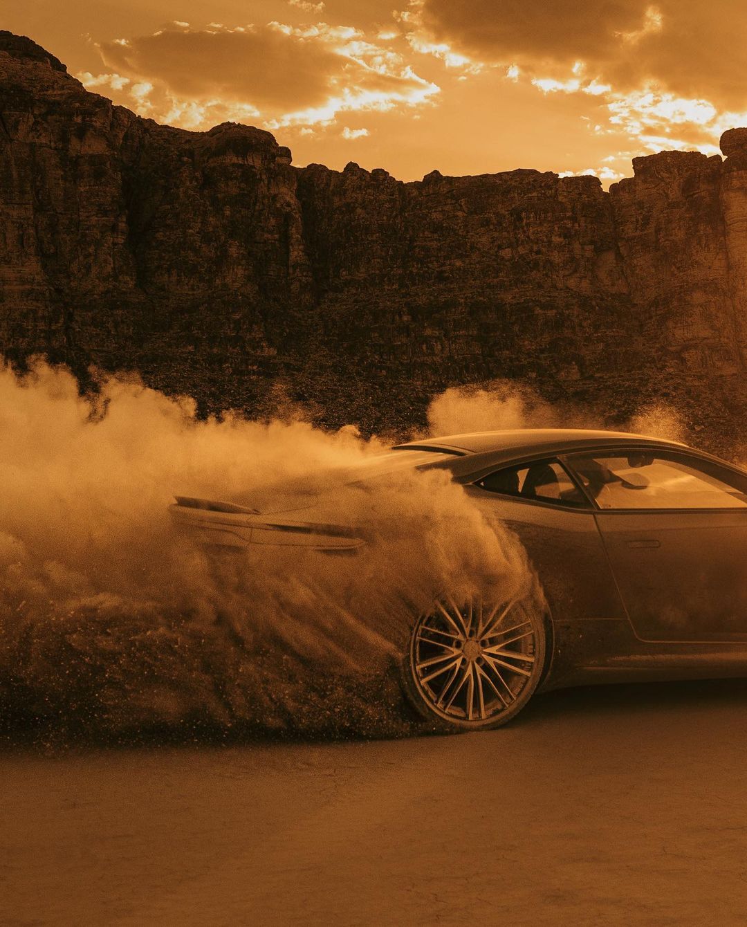 Automotive shot of a grey sports car drifting in a desert landscape with rock formations in the background, orange and sepia color grading.