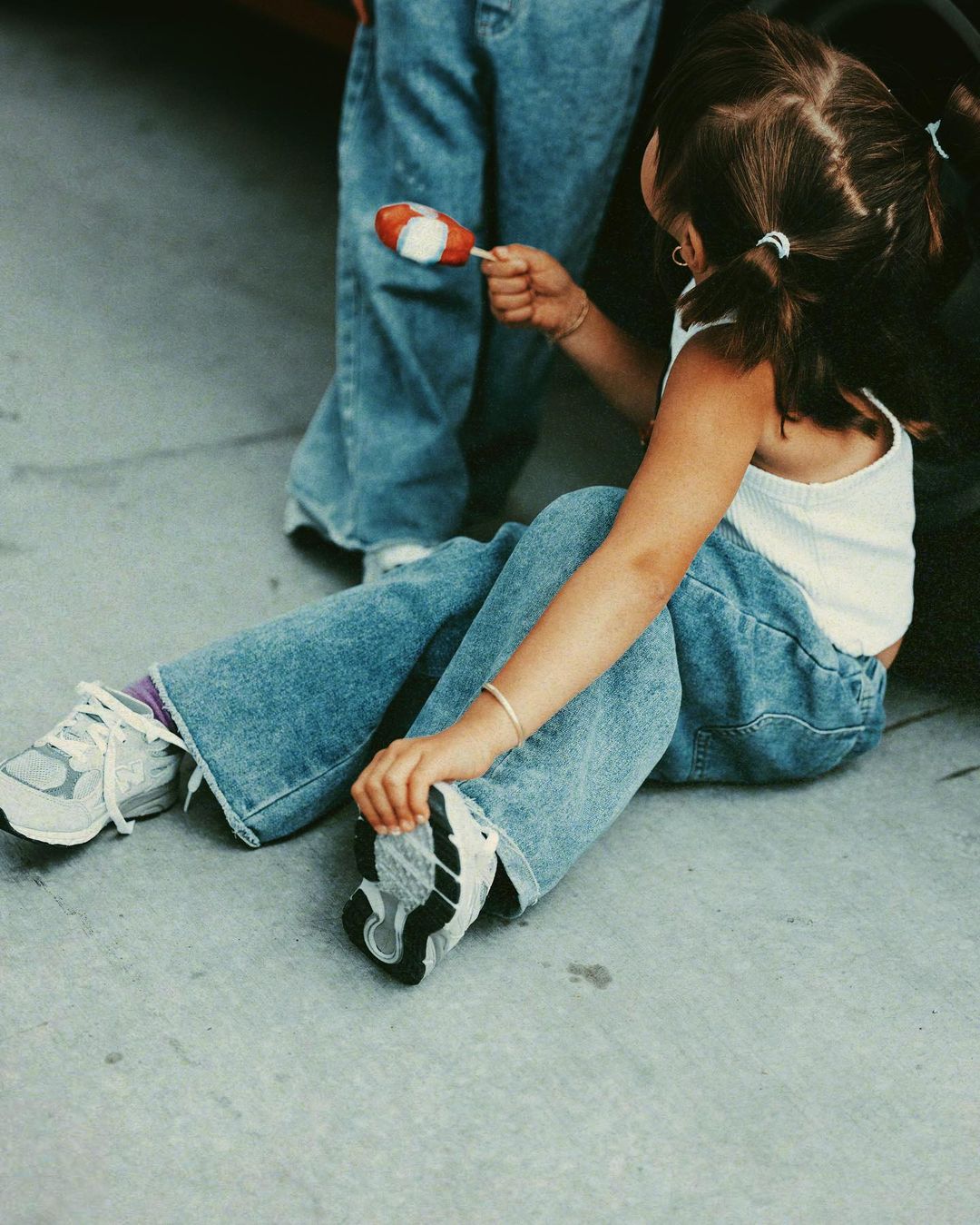 Young girl sits on the ground, holding a lollipop and adjusting her shoe, while another person stands nearby wearing jeans.