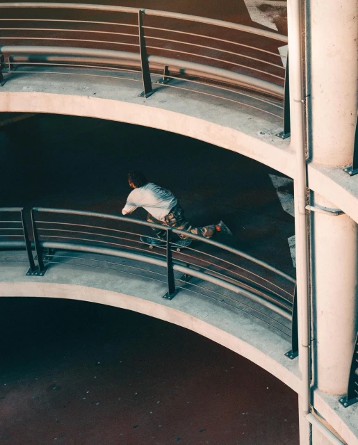 Adult rides a skateboard across a concrete parking ramp with metal railings and support columns in an urban environment.