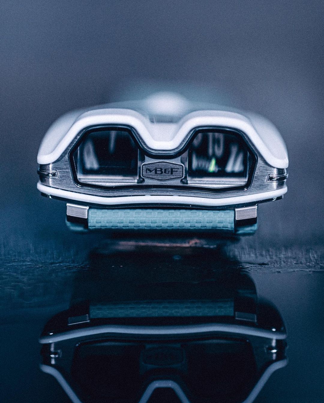 MB&F Horological Machine N°9 'Flow' wristwatch close-up with a white and metal case, displayed on a reflective surface, studio lit.