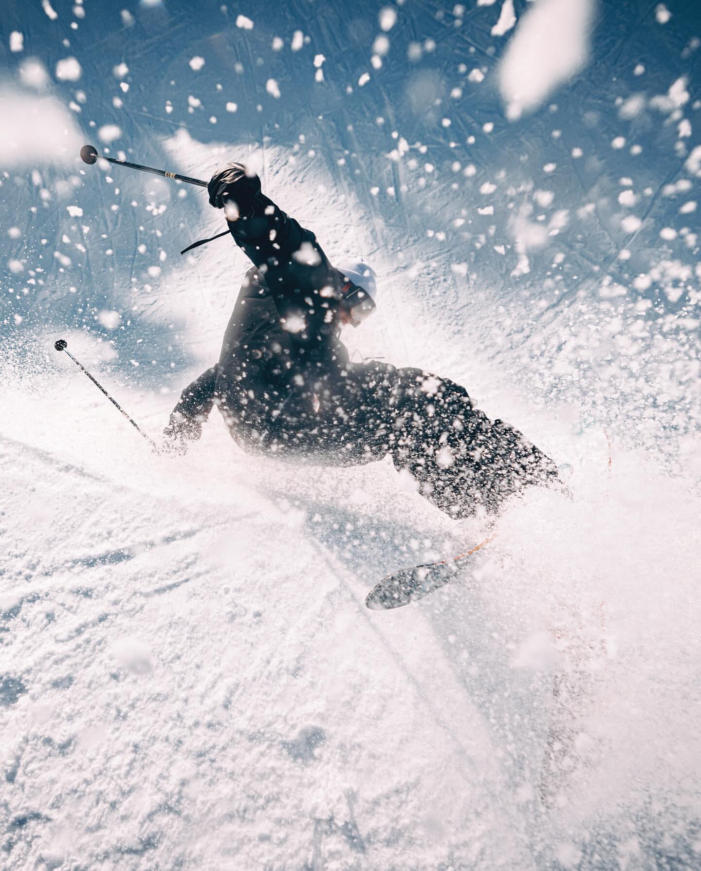 A person skis down a snow-covered slope against a blue sky. The skier is surrounded by a cloud of snow, captured from a low angle.