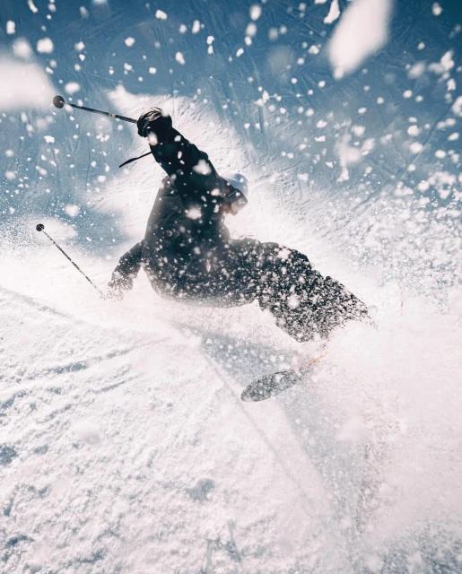 A person skis down a snow-covered slope against a blue sky. The skier is surrounded by a cloud of snow, captured from a low angle.