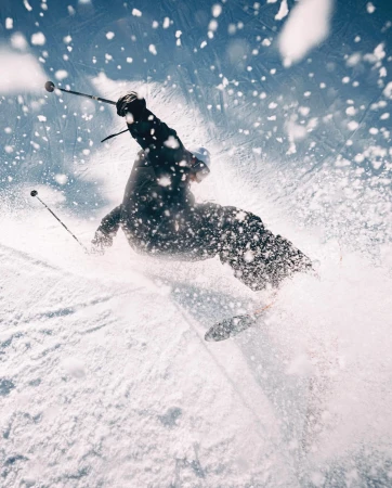 A person skis down a snow-covered slope against a blue sky. The skier is surrounded by a cloud of snow, captured from a low angle.