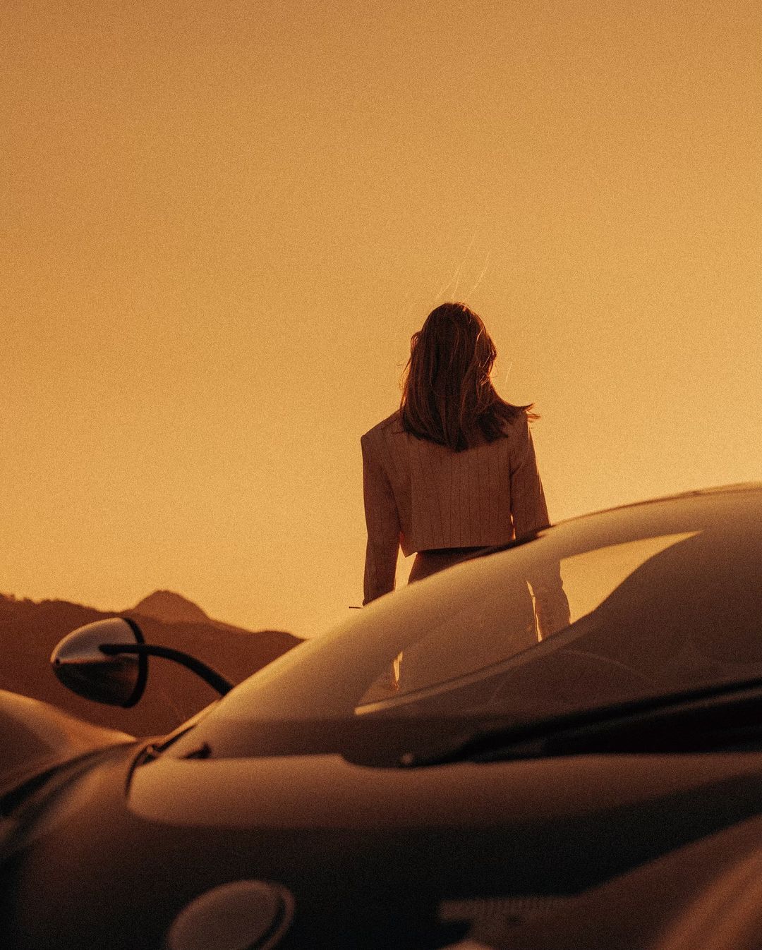 Woman standing near a sports car in a desert landscape, bathed in warm, golden light. Mountains in the background, automotive scene.