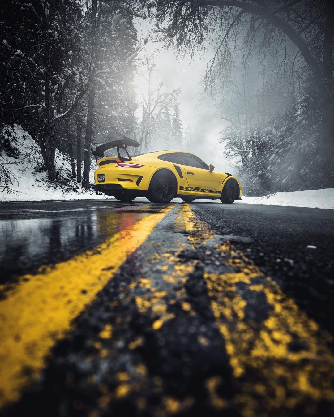 Yellow Porsche sports car parked on a snowy road lined with snow-covered trees in winter. The road has yellow lines painted on it.