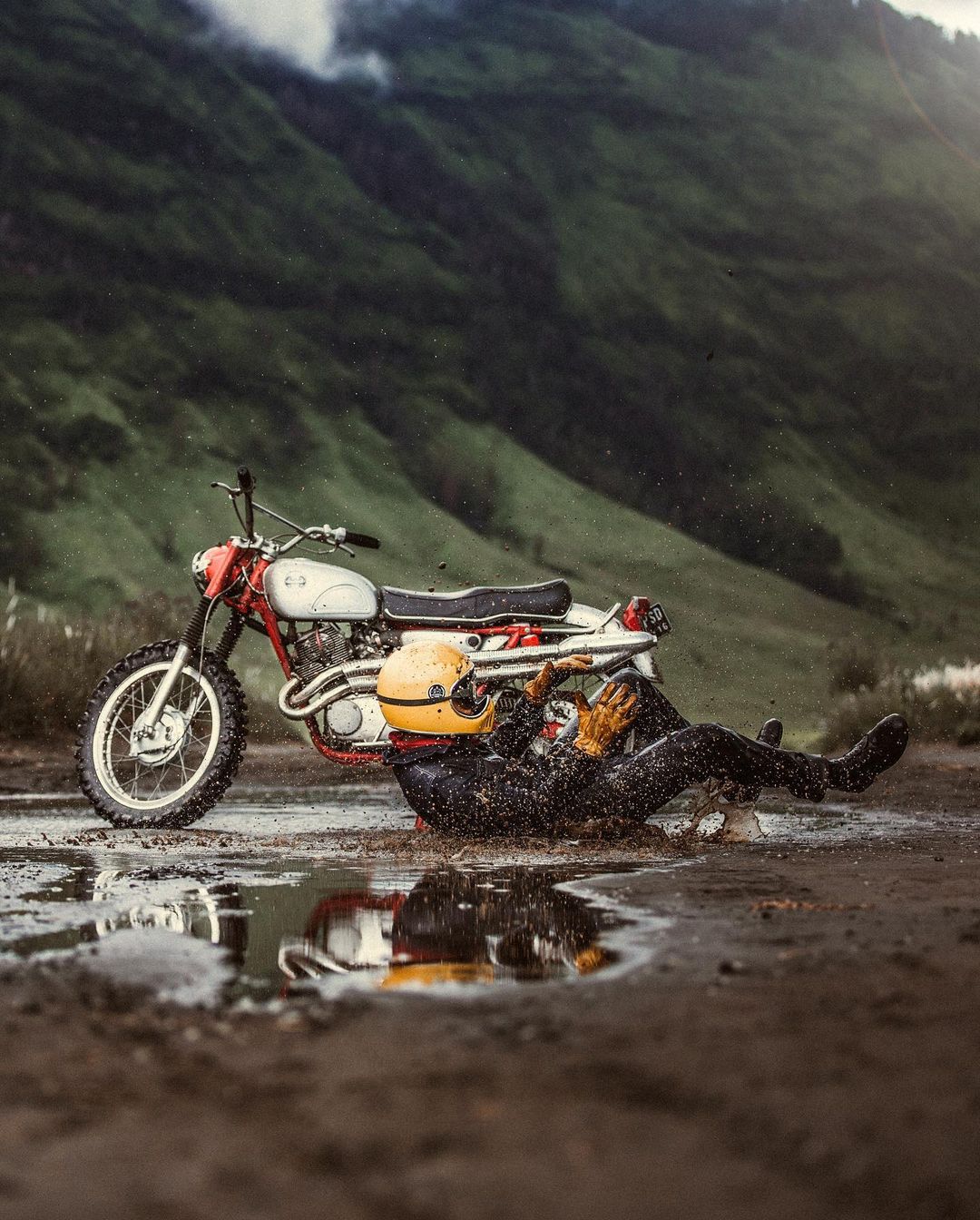 Motorcycle lying on its side in a muddy puddle with a fallen rider, mountains and cloudy sky in the background. Motorcycle helmet nearby.