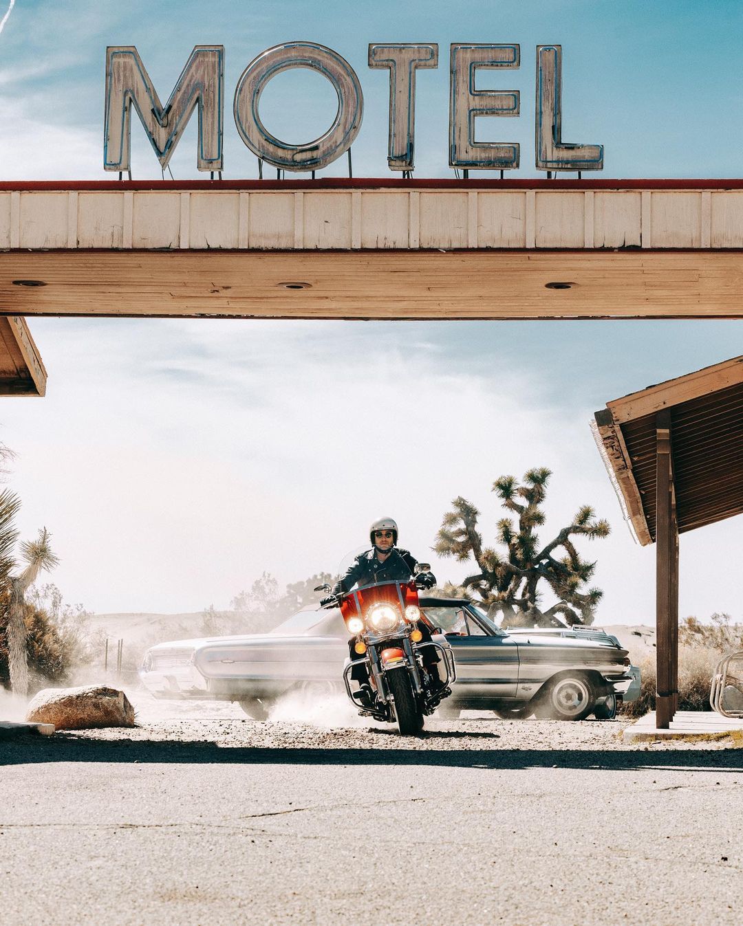Motorcyclist on a red motorcycle in a desert landscape with a vintage car and a retro motel sign on a sunny day. 