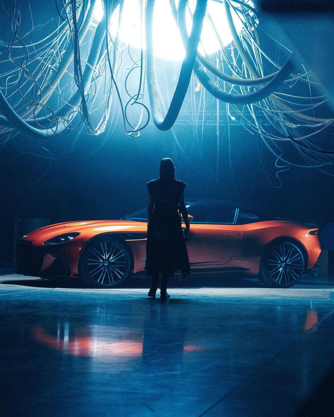 Profile of an adult woman in dark clothing with her back to the camera looking at an orange sports car under hanging cables.
