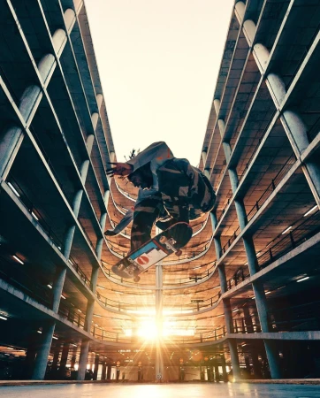 An urban action shot capturing a skateboarder performing a trick mid-air within a parking garage under bright sunlight, creating a dynamic scene.