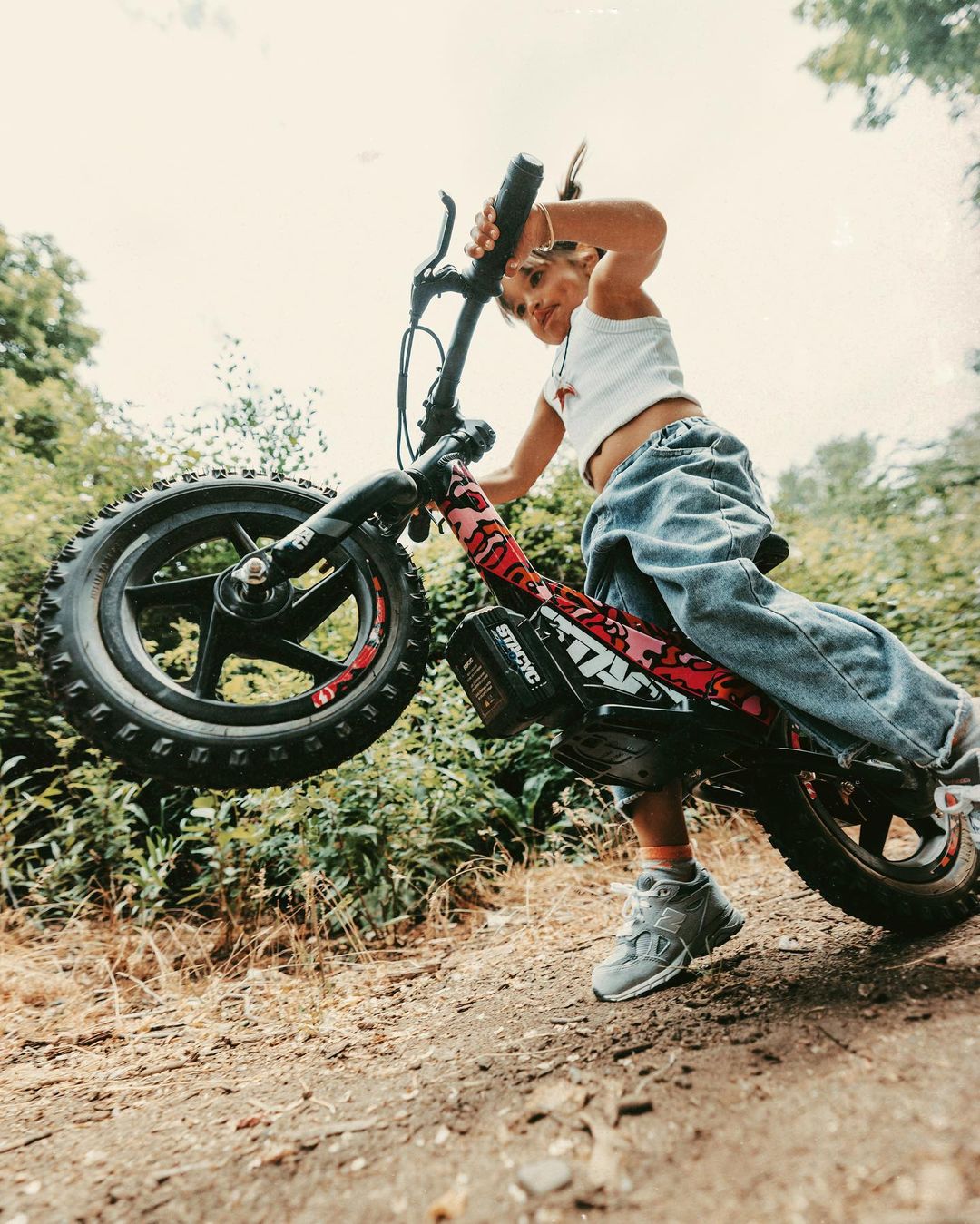 Child on a Strider STACYC electric balance bike performs a wheelie on a dirt path surrounded by green foliage under an overcast sky.