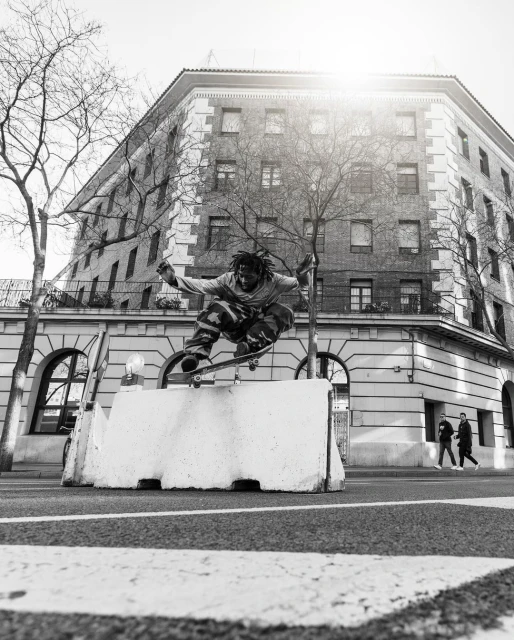 Monochrome action shot of a skateboarder performing a trick over a concrete barrier in an urban setting, building and pedestrians in the background.