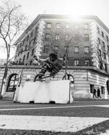 Black man performing a skateboard jump over a concrete road barrier with a multi-story building in the background