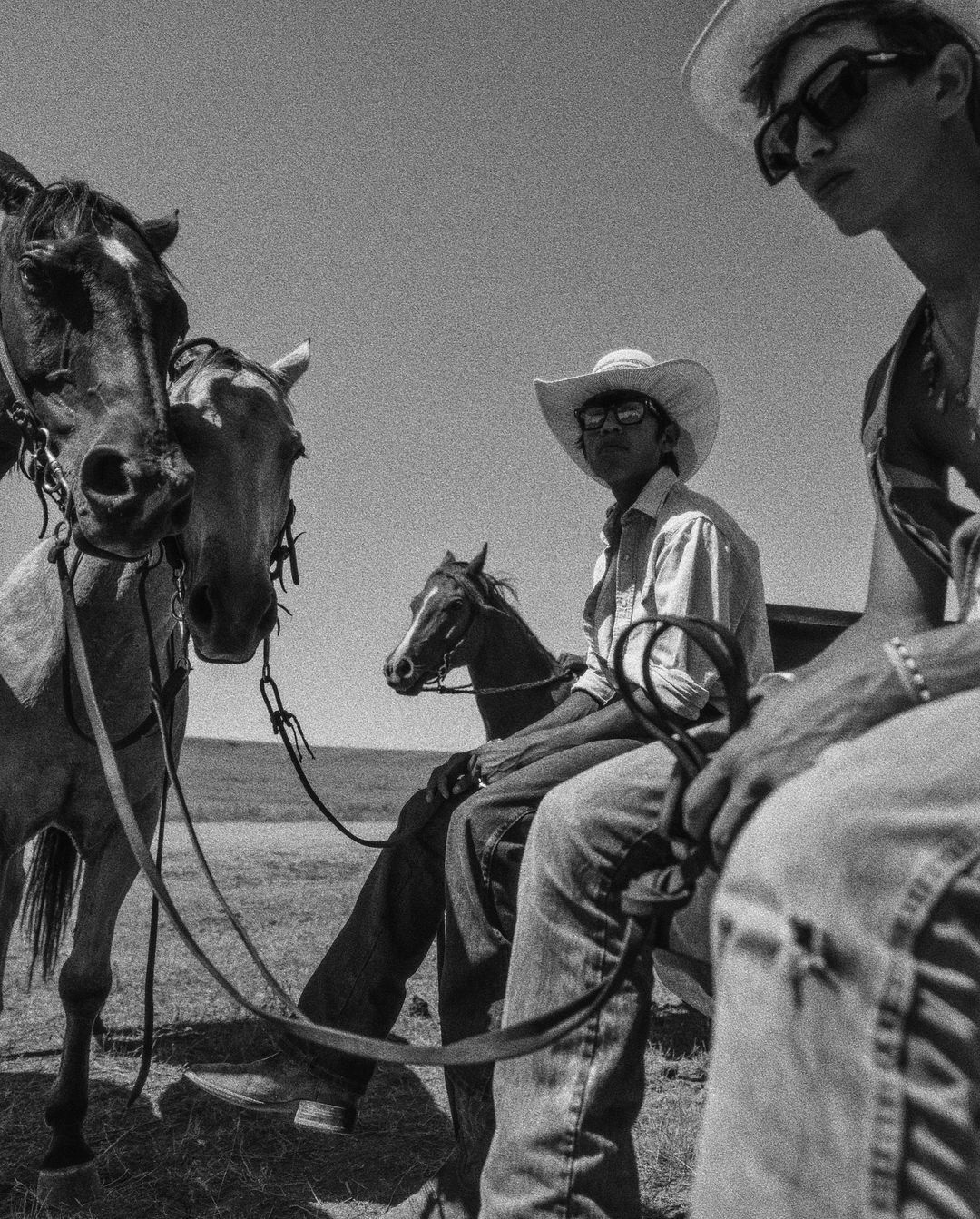 Black and white photo of two men wearing cowboy hats next to three horses in a grassy outdoor environment on a bright day.