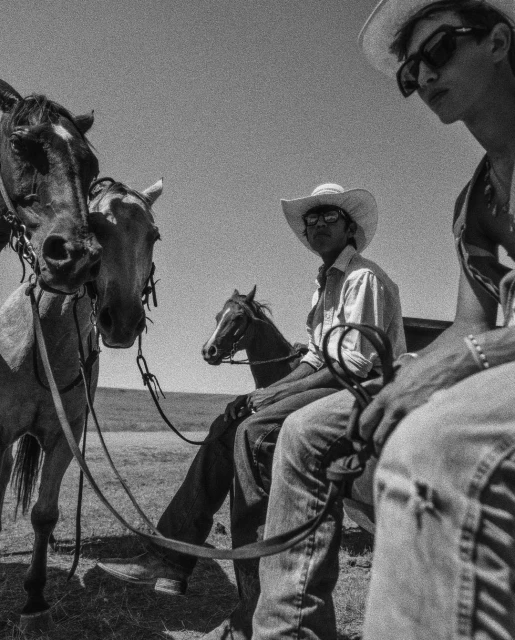 Black and white photo of two men wearing cowboy hats next to three horses in a grassy outdoor environment on a bright day.