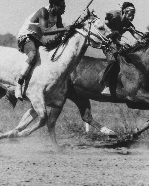 Two men on horseback racing, with one rider wearing a headband, captured in a vintage black and white photograph on a dusty course.