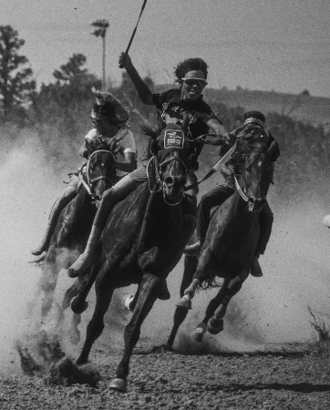 Three horses and jockeys racing down a dirt track, kicking up dust on a sunny day, captured in a monochrome action shot.