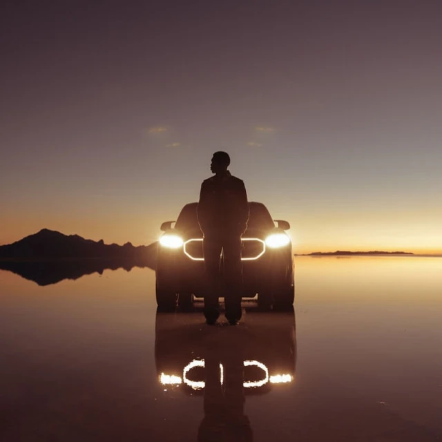 A man standing in front of a car on a flat, reflective surface, possibly water, with a colorful sunset and mountains in the background.
