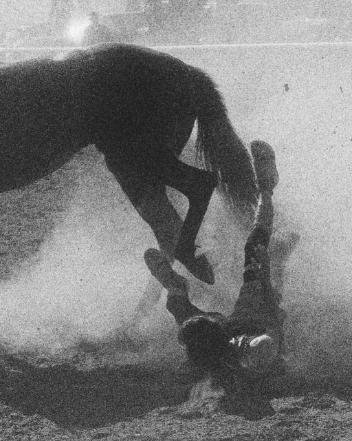 Black and white photo captures a rodeo scene with a rider falling from a horse in a dusty arena, emphasizing the action and potential danger.