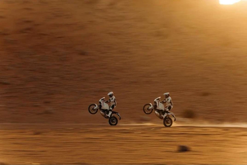 Two motorcyclists perform a wheelie on dirt bikes, captured in a wide outdoor shot against a background of a desert landscape.