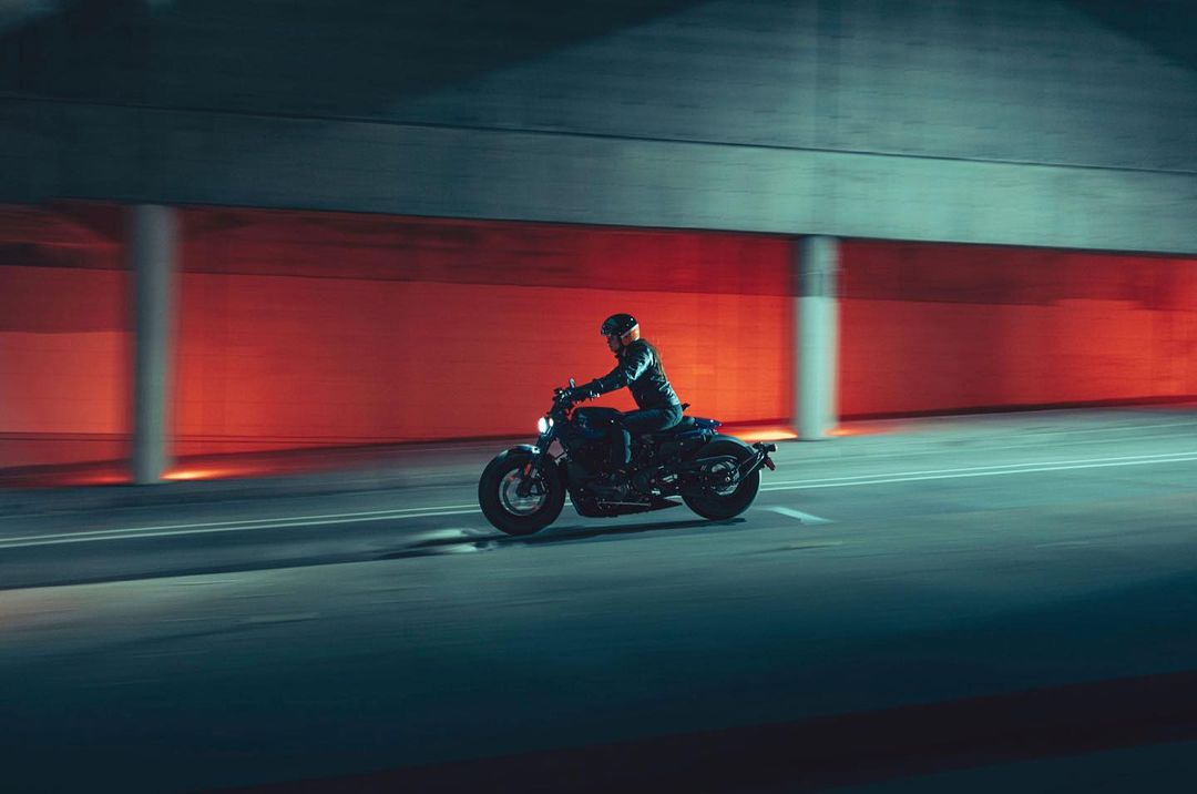 A motorcyclist rides a black motorcycle at night through a concrete tunnel with diffused red lighting in the background.