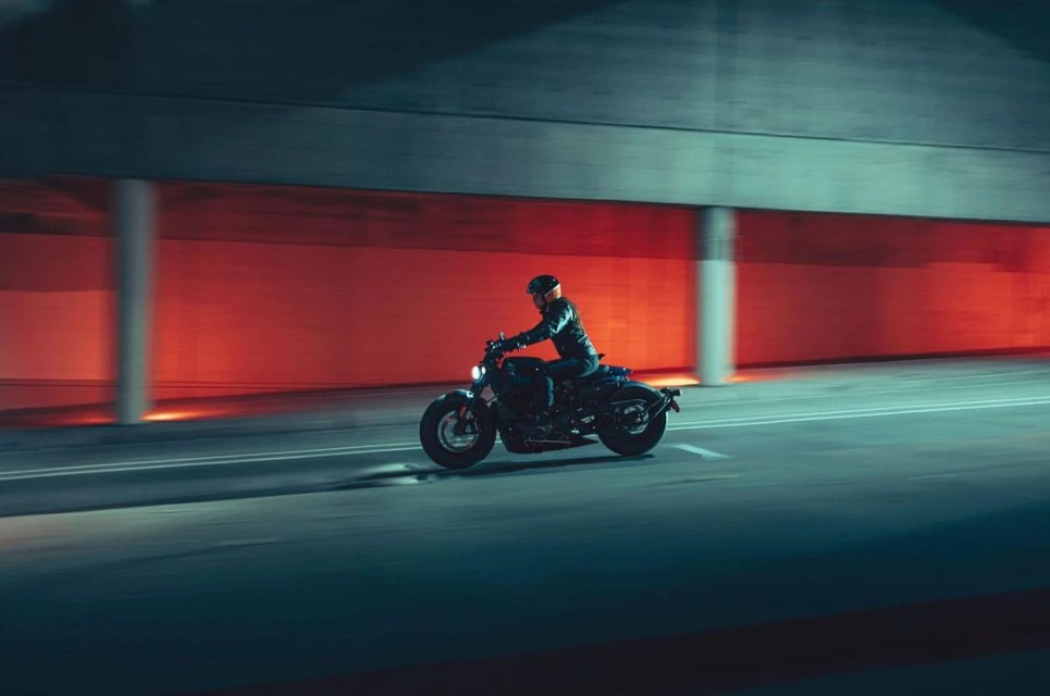 A motorcyclist rides a black motorcycle at night through a concrete tunnel with diffused red lighting in the background.