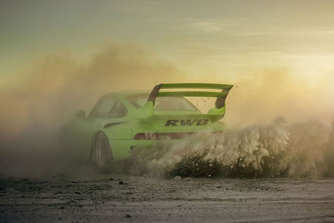 A lime-green Porsche 911 drifts powerfully in a dusty outdoor setting, creating a cloud of dirt with motion blur in the landscape.